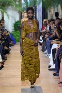 Model walks the runway in a mustard crochet two-piece outfit with a matching crochet bag, flanked by seated spectators taking photos.