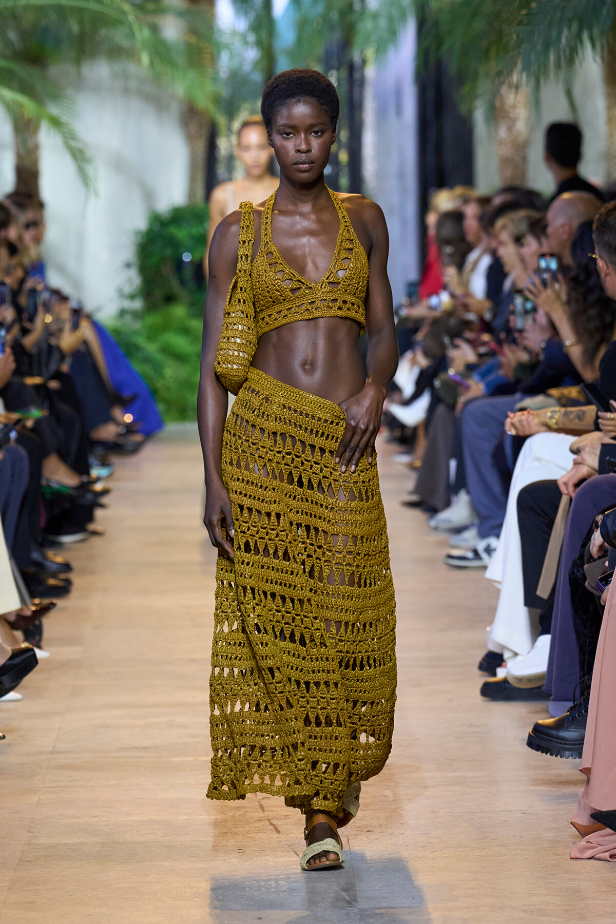 Model walks the runway in a mustard crochet two-piece outfit with a matching crochet bag, flanked by seated spectators taking photos.
