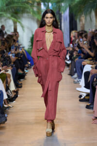 Model walks runway in dusty-rose utility jumpsuit with large front pockets and deep V-neck, gold necklace, beige platform sandals; audience watches.