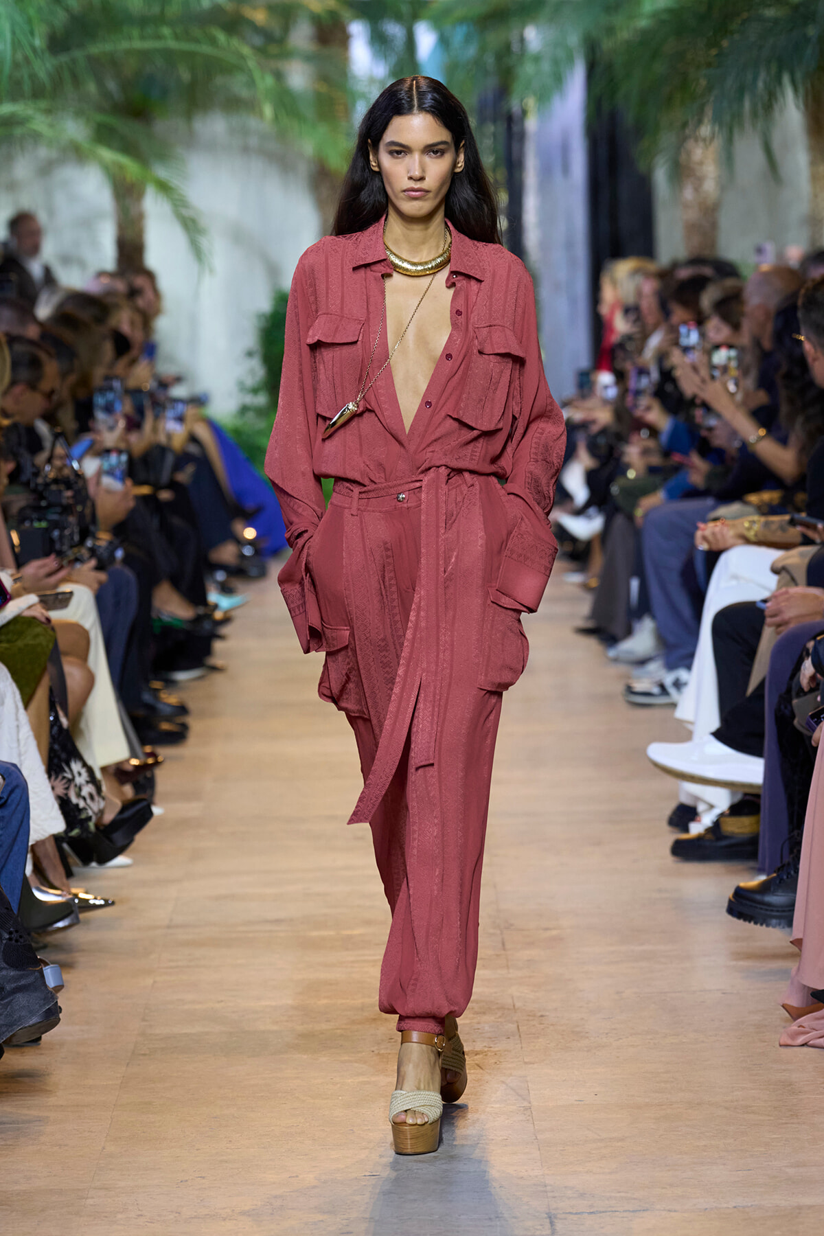 Model walks runway in dusty-rose utility jumpsuit with large front pockets and deep V-neck, gold necklace, beige platform sandals; audience watches.