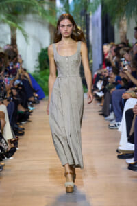 Model walks a fashion runway in a light gray sleeveless midi dress with embroidered bodice and seam detailing, audience flanking the catwalk.