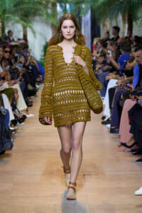 Model walks the runway in a mustard crochet dress with matching bag, audience on both sides.