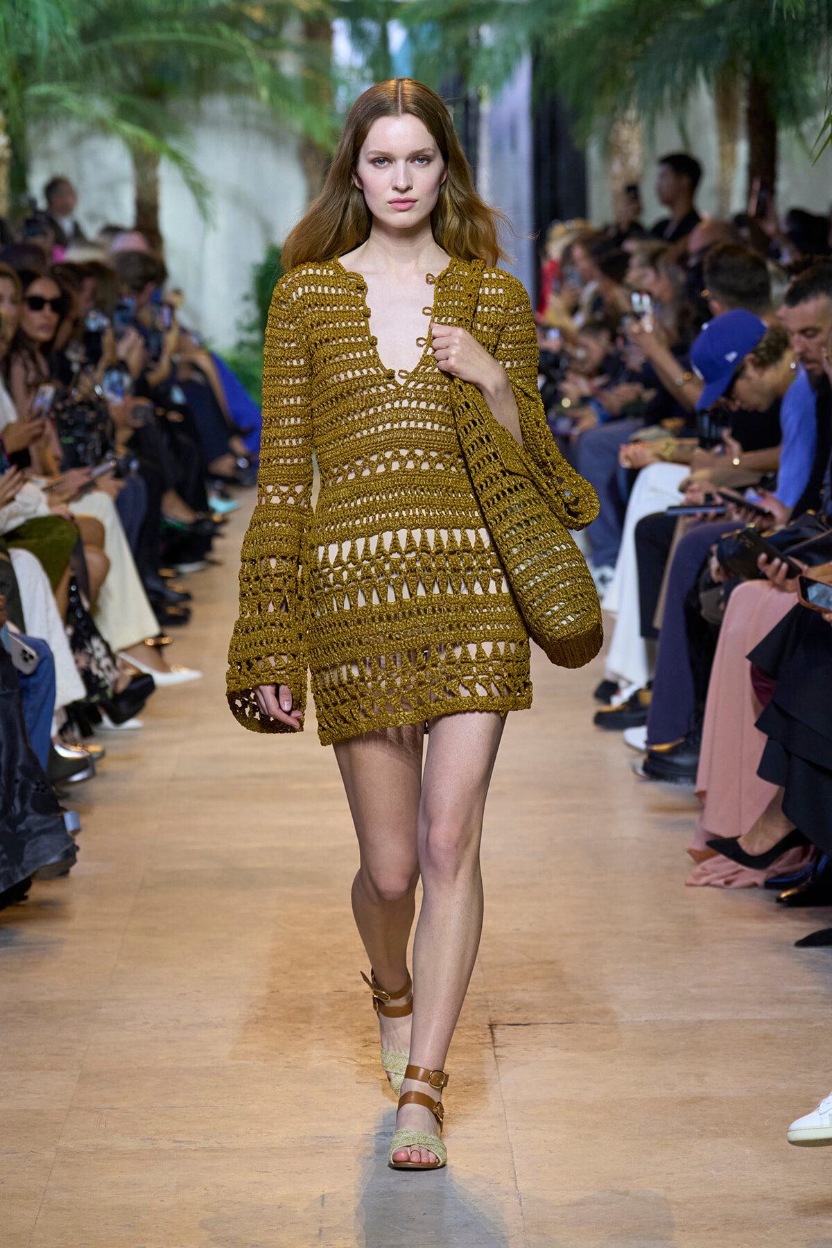 Model walks the runway in a mustard crochet dress with matching bag, audience on both sides.