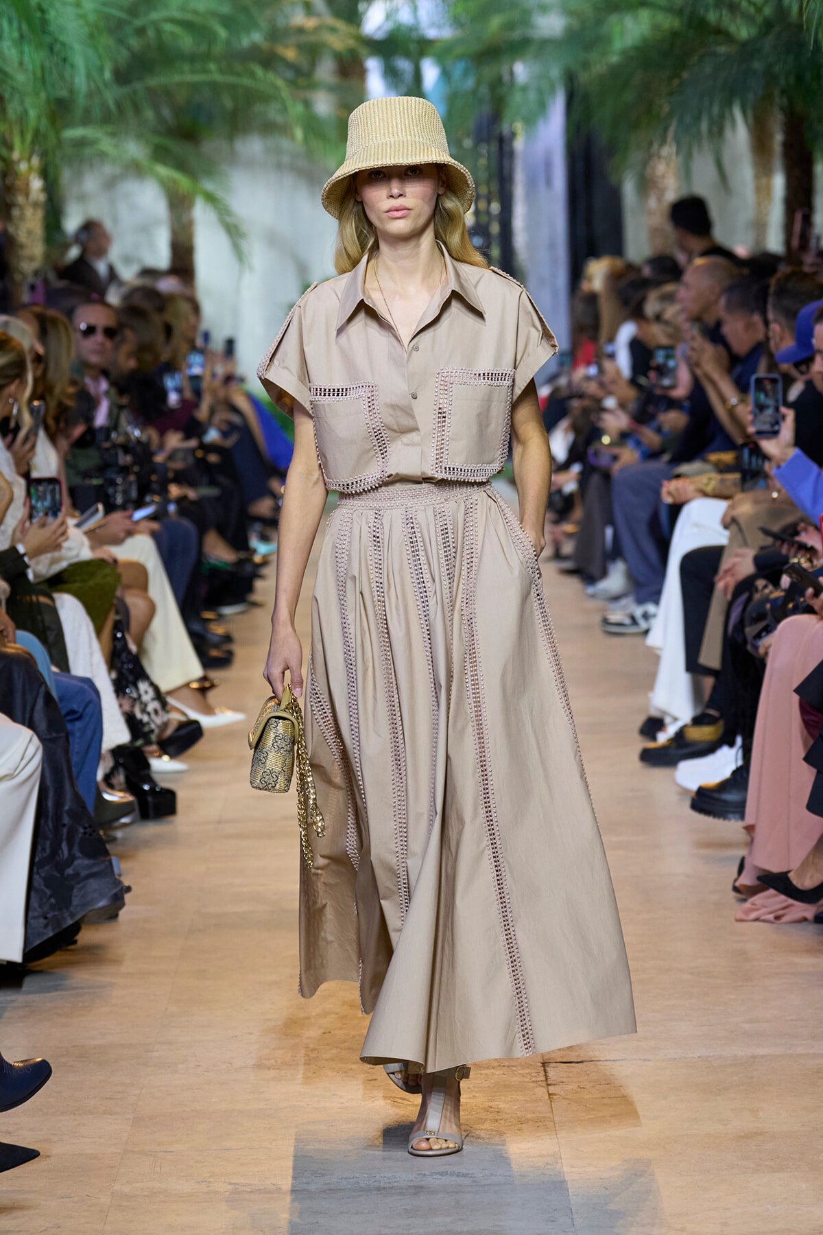 Model walks the runway in a beige wide-leg dress with stitched trim and two large front pockets, paired with a beige straw hat and small patterned handbag on the arm.