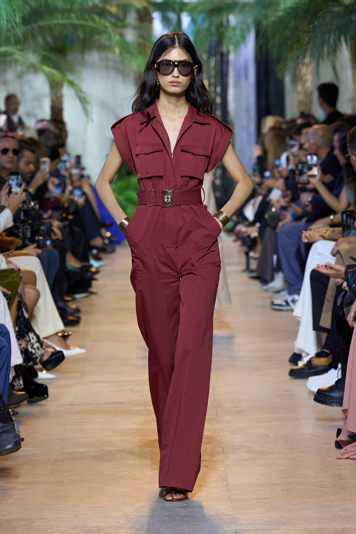 Model in burgundy sleeveless jumpsuit with a wide belt walks a fashion runway, wearing dark sunglasses.