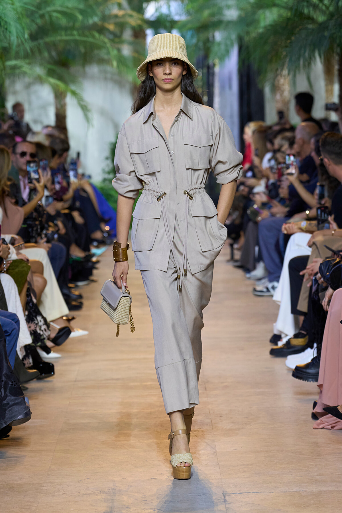 Model walking a beige utility jumpsuit with a cinched waist on a fashion runway, wearing a straw hat and carrying a small beige handbag.