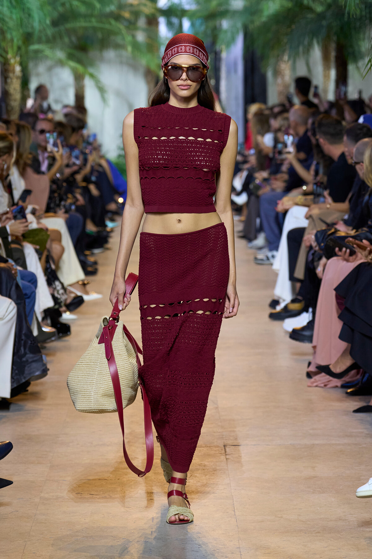 Model walks a runway wearing a burgundy crochet crop top and matching skirt, oversized sunglasses, and a red headscarf with a straw bag.