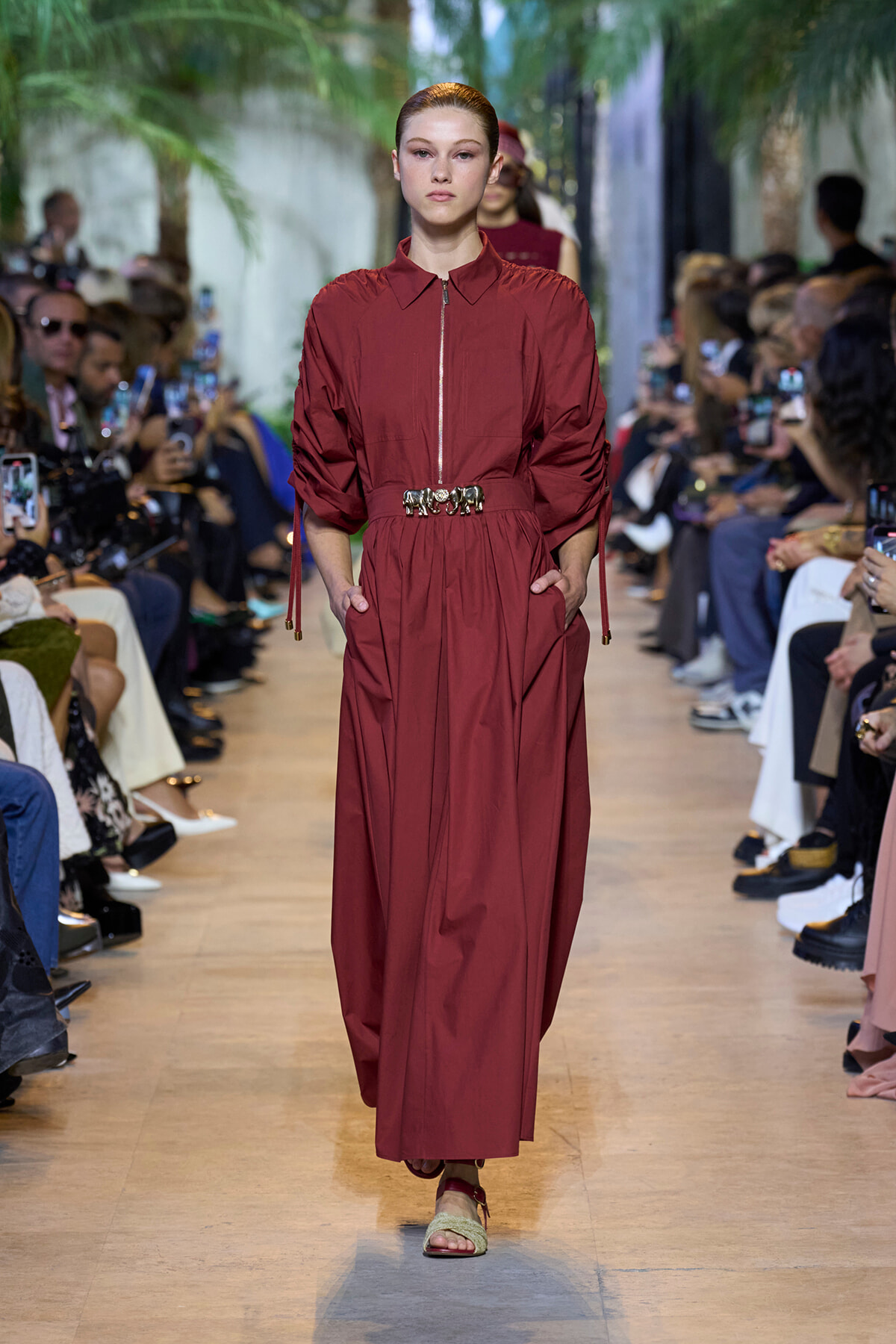 Model walks a fashion runway in a burgundy, long-sleeve zip-front dress with a gathered waist and a metallic belt, flanked by paparazzi and spectators.