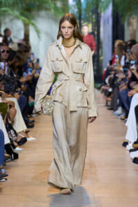 Model walking a runway in a beige belted utility jumpsuit with large pockets, gold necklace, and patterned clutch