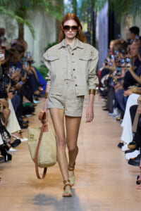 Model walks the runway in a beige denim jacket and matching shorts, oversized sunglasses, and a large straw tote.