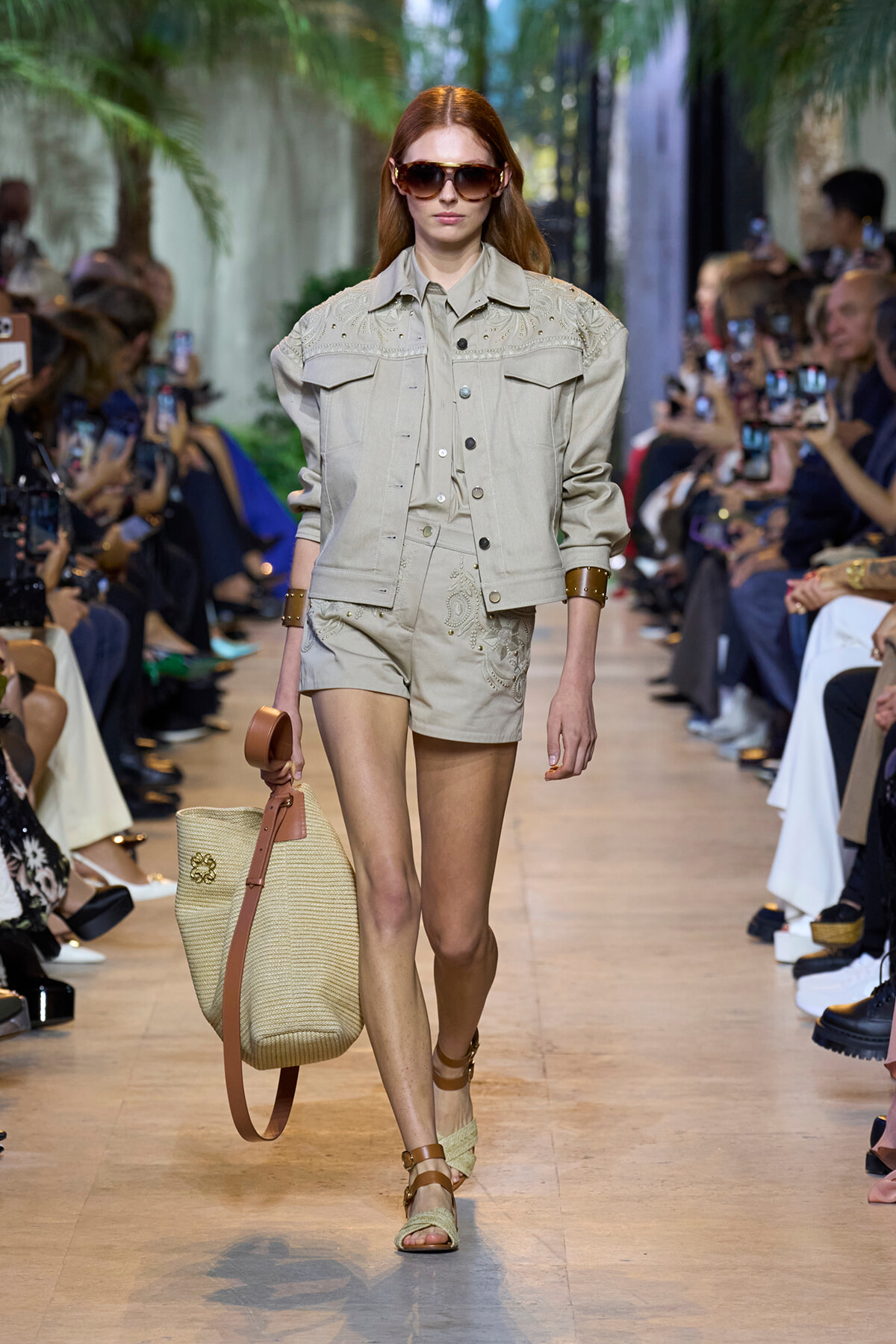 Model walks the runway in a beige denim jacket and matching shorts, oversized sunglasses, and a large straw tote.
