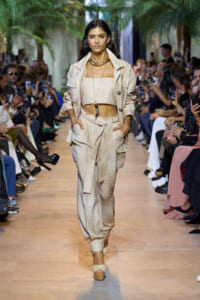 Model walks a runway in a beige coordinated suit with a cropped top, belt, and layered necklace; audience filming on both sides.