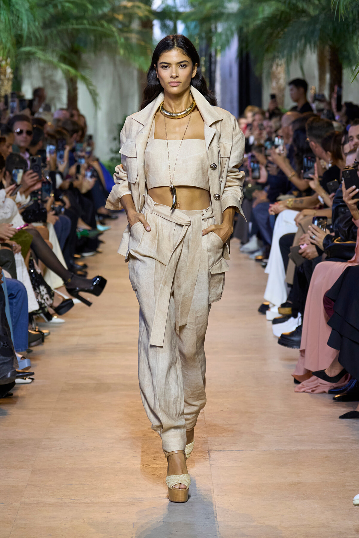 Model walks a runway in a beige coordinated suit with a cropped top, belt, and layered necklace; audience filming on both sides.