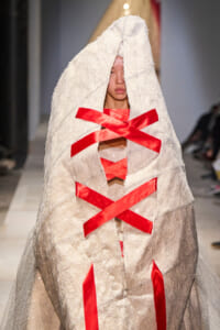 Model on a runway wearing a metallic silver cloak with a hood and bold red satin ribbon crisscross lacing across the front, obscuring the face partially.