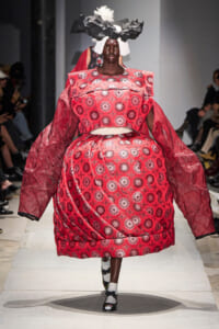 Model walks a runway in an oversized red dress with white circular patterns and dramatic puff sleeves, wearing a sculptural black-and-white headpiece.