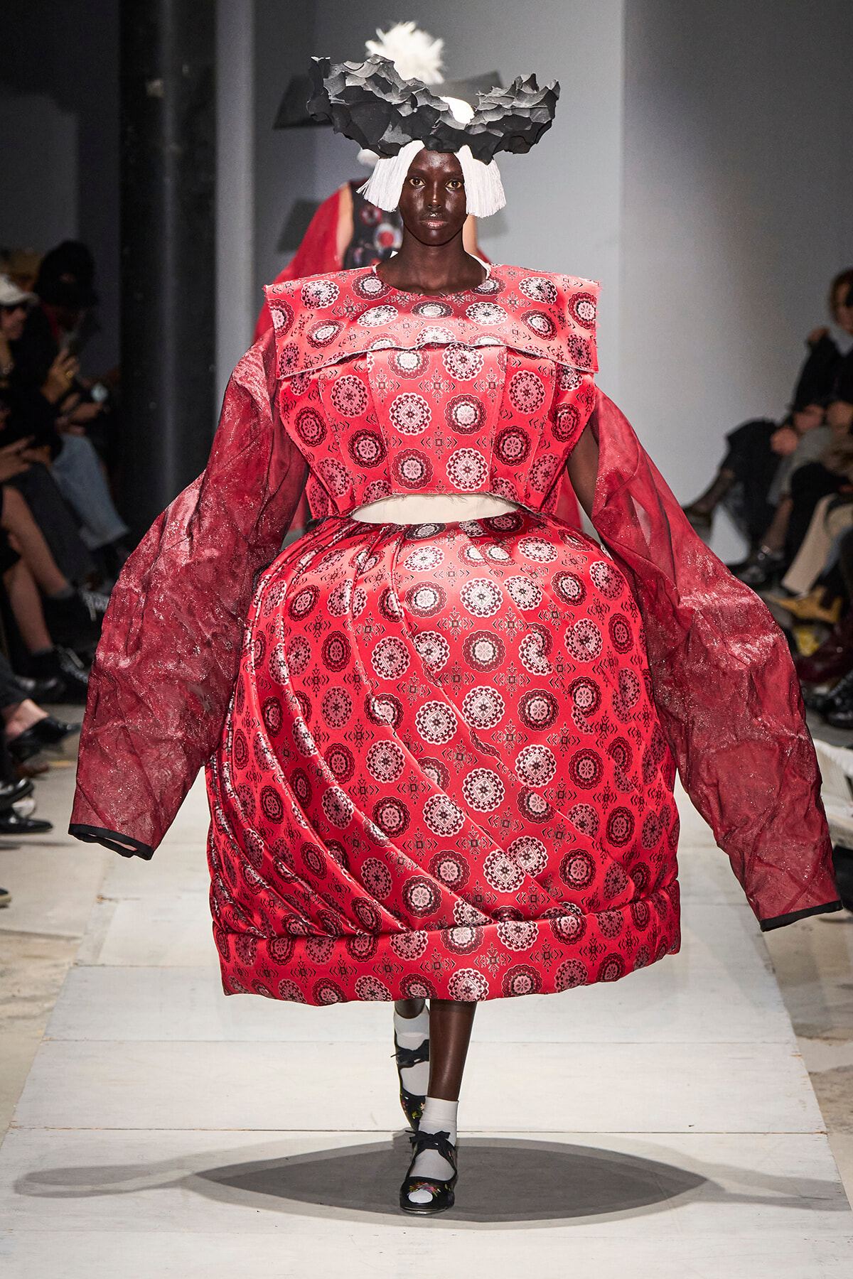 Model walks a runway in an oversized red dress with white circular patterns and dramatic puff sleeves, wearing a sculptural black-and-white headpiece.