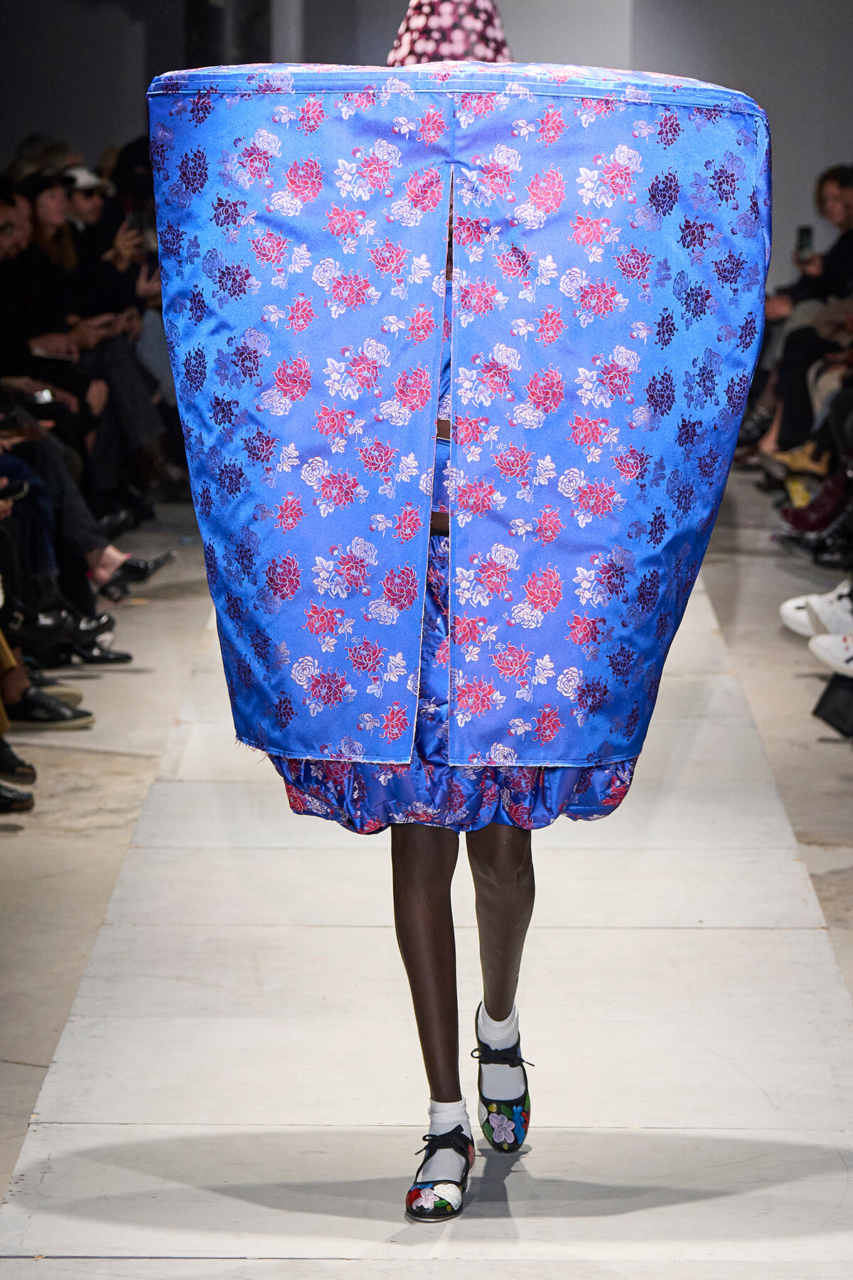Model on a runway wearing a voluminous blue dress covered in pink and white floral print; the garment obscures the torso and extends to the knees, revealing the model's legs and floral shoes.