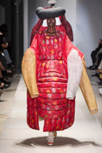 Model on a runway wearing a bold red floral quilted gown with oversized gold and white arm panels and a large sculptural black hat on a white catwalk.