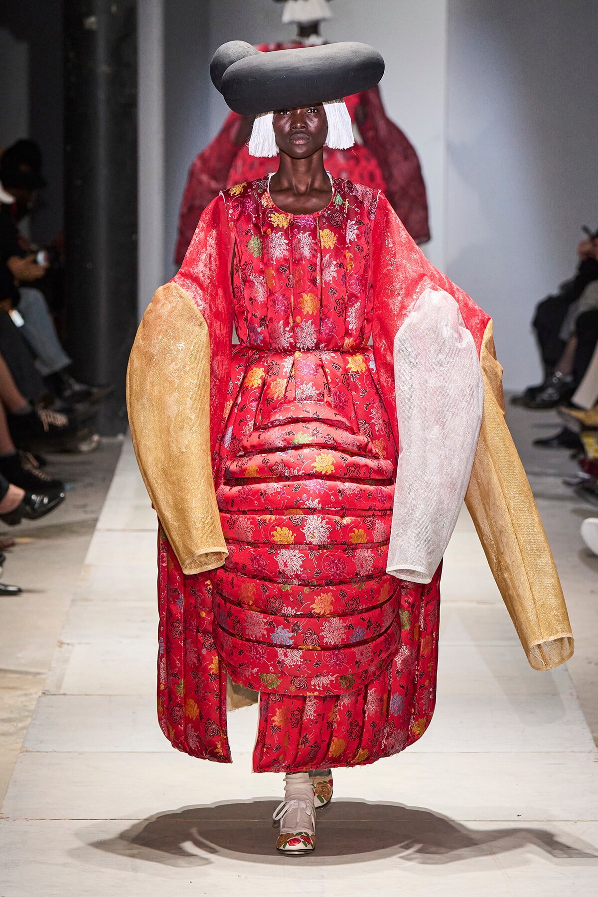 Model on a runway wearing a bold red floral quilted gown with oversized gold and white arm panels and a large sculptural black hat on a white catwalk.