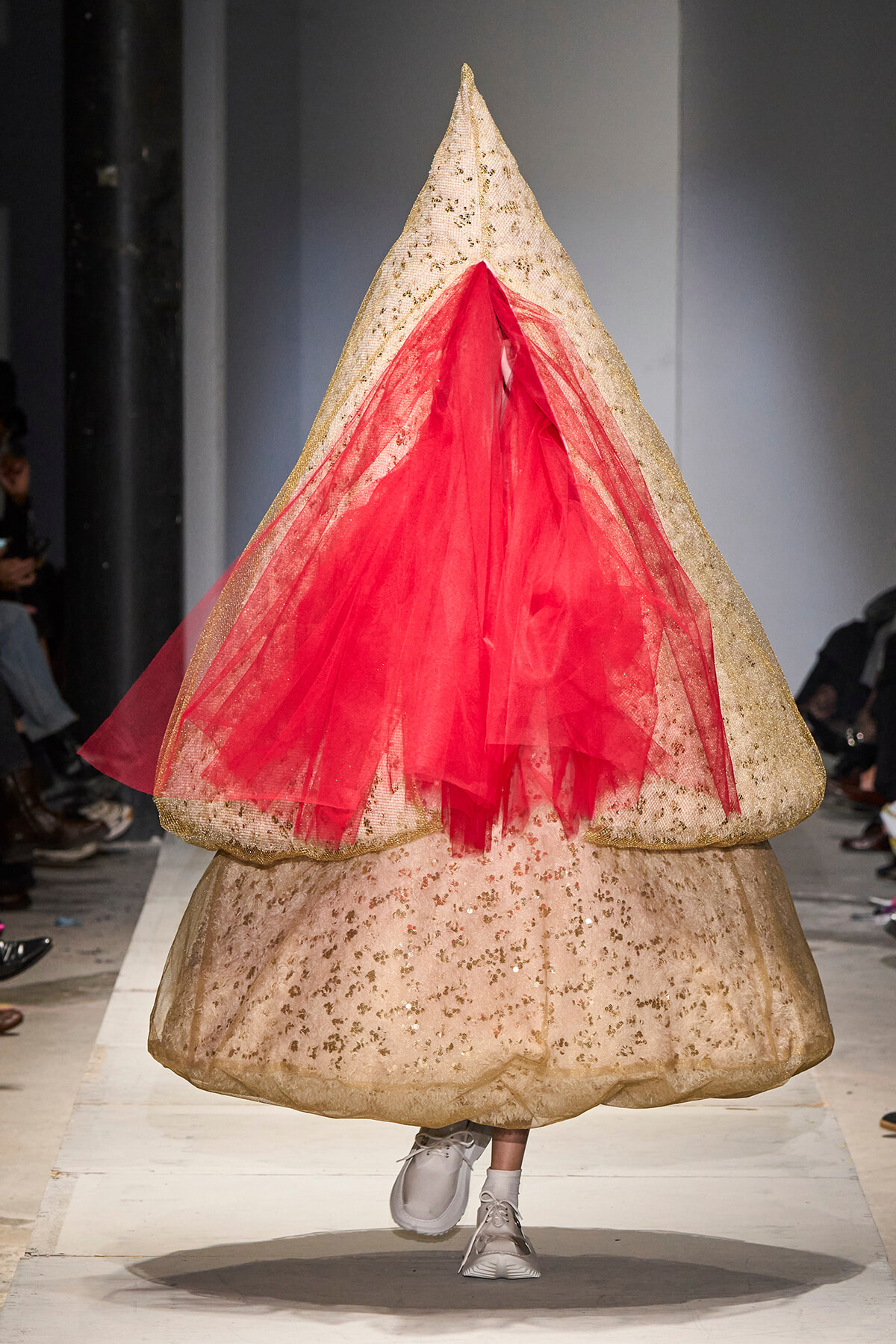 Model on a white runway wearing a giant beige sequined cone gown with red tulle draped at the front.