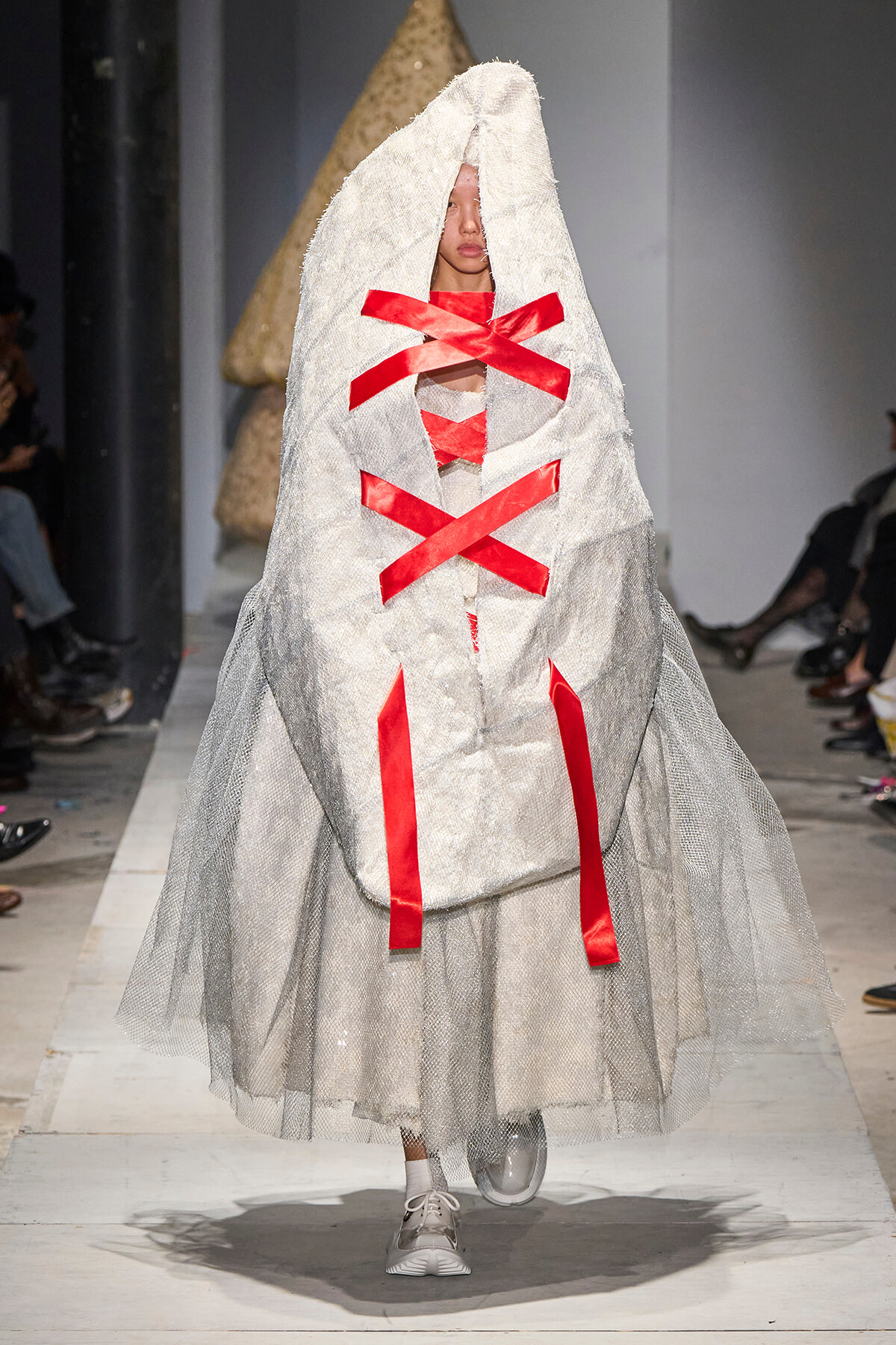 Model on a runway wearing an oversized gray hooded dress with red ribbon lacing in X shapes across the front and sheer tulle layering, paired with white sneakers.