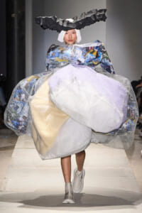 Model walking the runway in a dramatic sculptural gown made of layered mesh, plastic, and fabric with a jagged black headpiece.