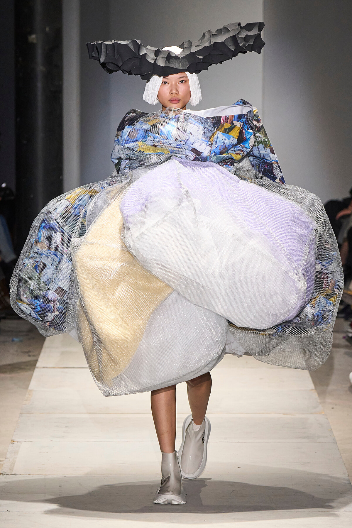 Model walking the runway in a dramatic sculptural gown made of layered mesh, plastic, and fabric with a jagged black headpiece.