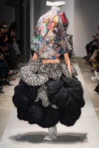 Runway model in an oversized black fluffy skirt with sculptural black-and-white patterned ruching and a colorful collage-printed hooded top; white sneakers.