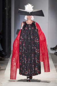 Runway model in a black printed dress with red sheer cape sleeves and a large black angular hat with a white feathered top.