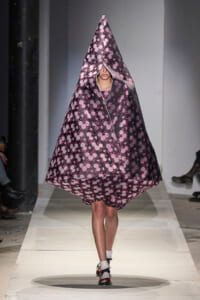 Model walks the runway in a voluminous, cone-shaped poncho with pink floral pattern on a dark background, covering her head and body.