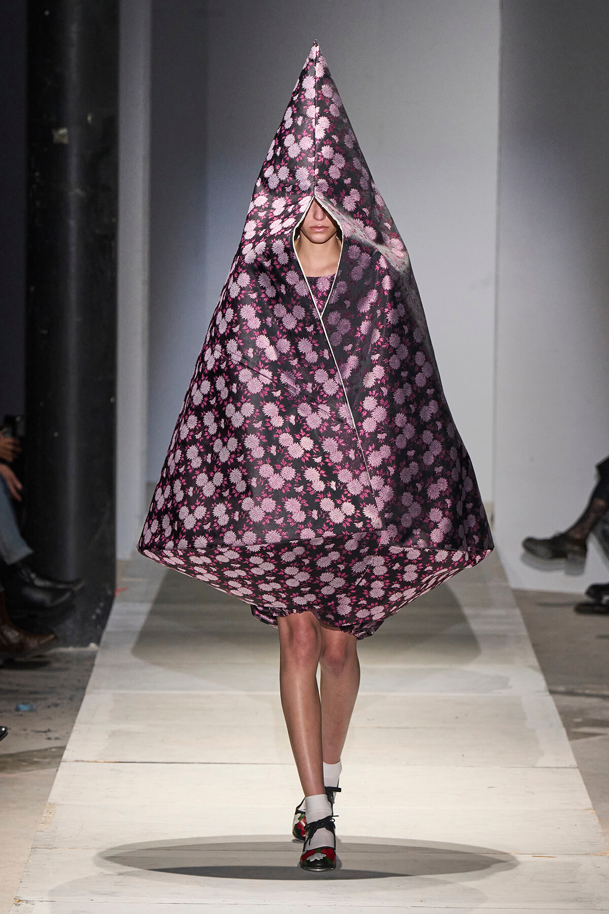 Model walks the runway in a voluminous, cone-shaped poncho with pink floral pattern on a dark background, covering her head and body.