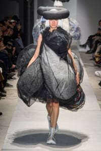 Model walks a runway in a dramatic metallic silver and black tulle dress with an oversized circular headpiece. Func.