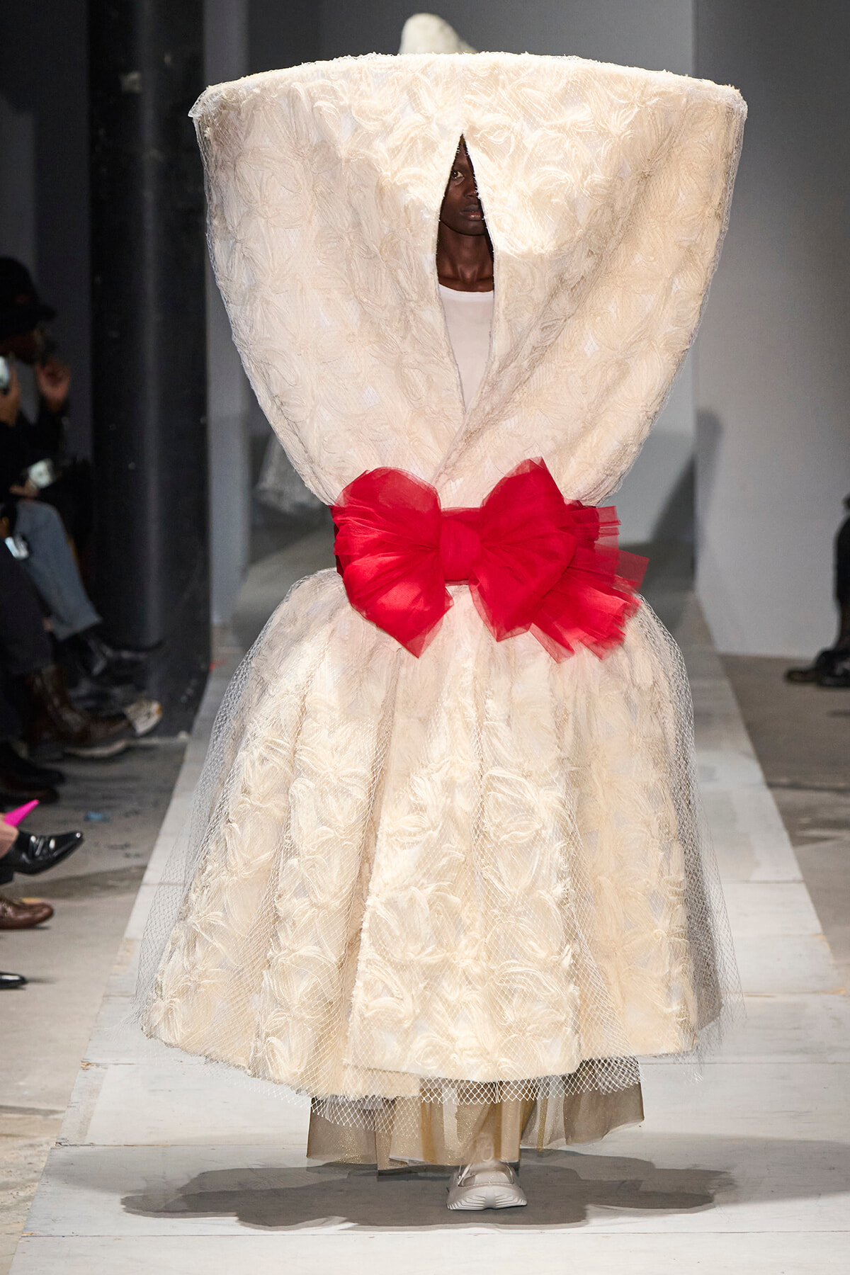 Model on a fashion runway wearing a voluminous ivory lace gown with a sculptural twisted bodice and a bright red ribbon bow at the waist, face peeking through a front opening.