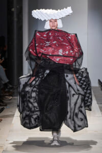 Runway model wearing an avant-garde red and black layered gown with sheer black overlay and a white sculptural headpiece, walking the catwalk.