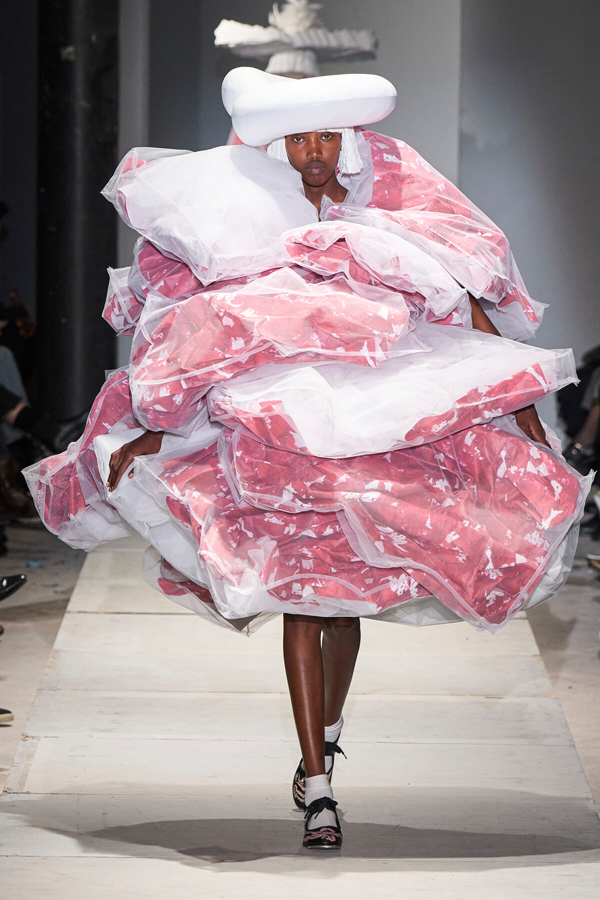 Model walks the runway in a voluminous pink and white sculptural gown with a white headpiece.
