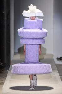 Model on runway wearing a tall lavender fuzzy sculpture dress made of stacked circular shapes, with a wide white hat and white boots