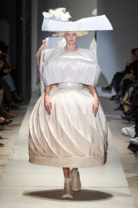 Model walking a runway in an oversized ivory-white gown with a puffed bodice and full, sculpted skirt, topped by a large angular hat.