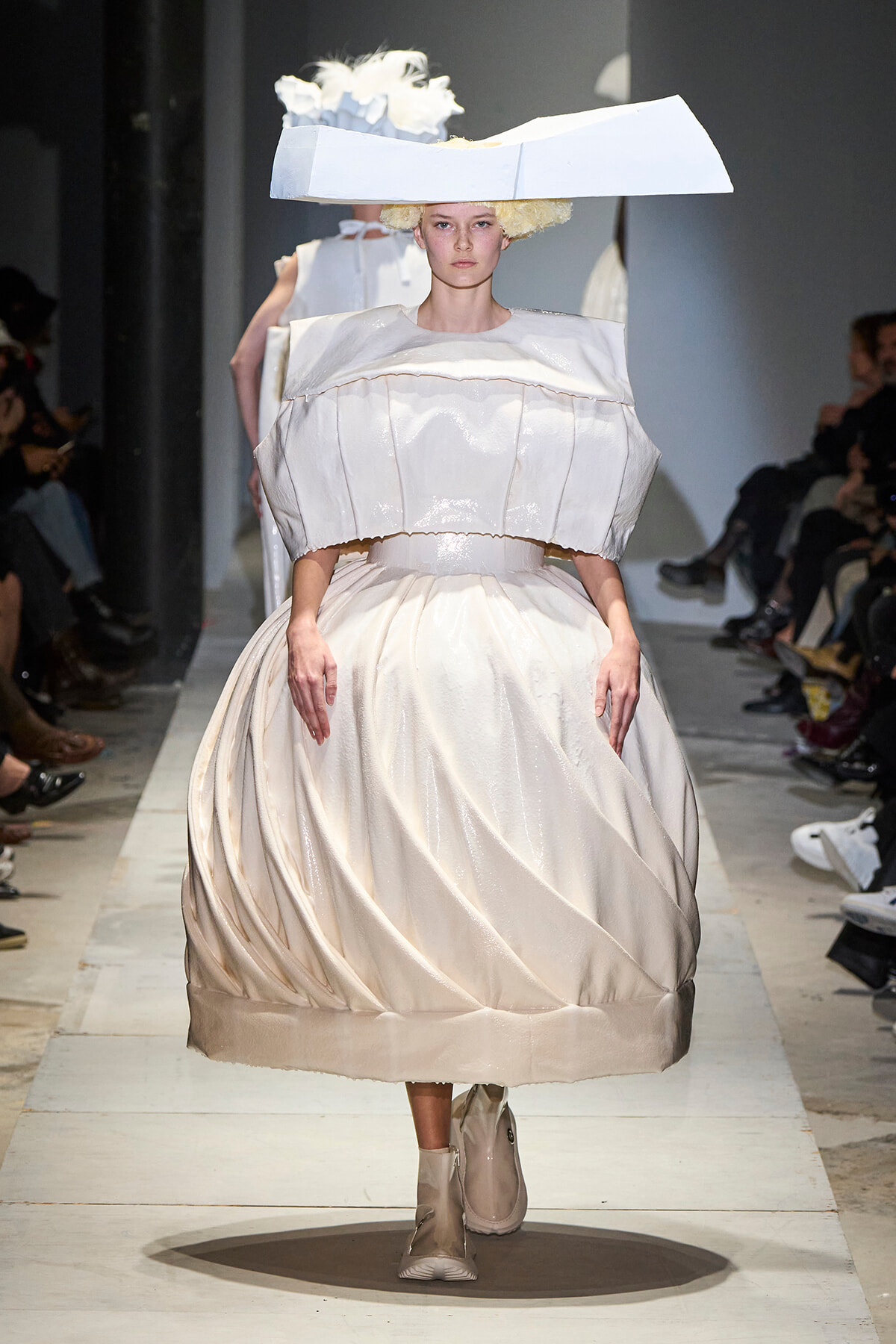 Model walking a runway in an oversized ivory-white gown with a puffed bodice and full, sculpted skirt, topped by a large angular hat.