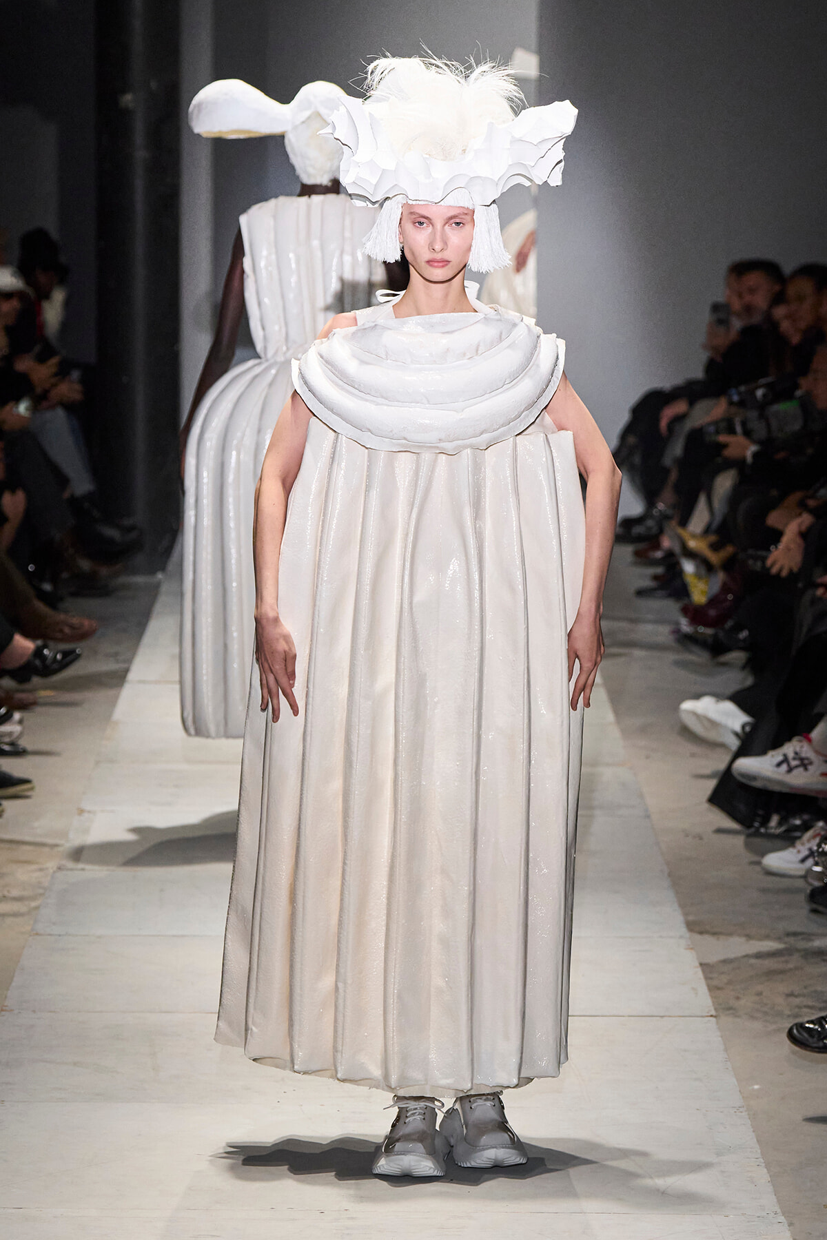 Model on a runway wearing a voluminous white gown with an oversized layered collar and a dramatic sculptural headpiece.