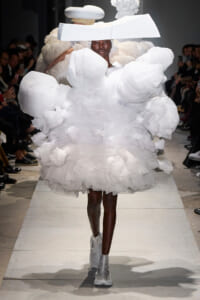 Model on a runway wearing a dramatic white sculptural dress with oversized ruffles and a wide white headpiece.