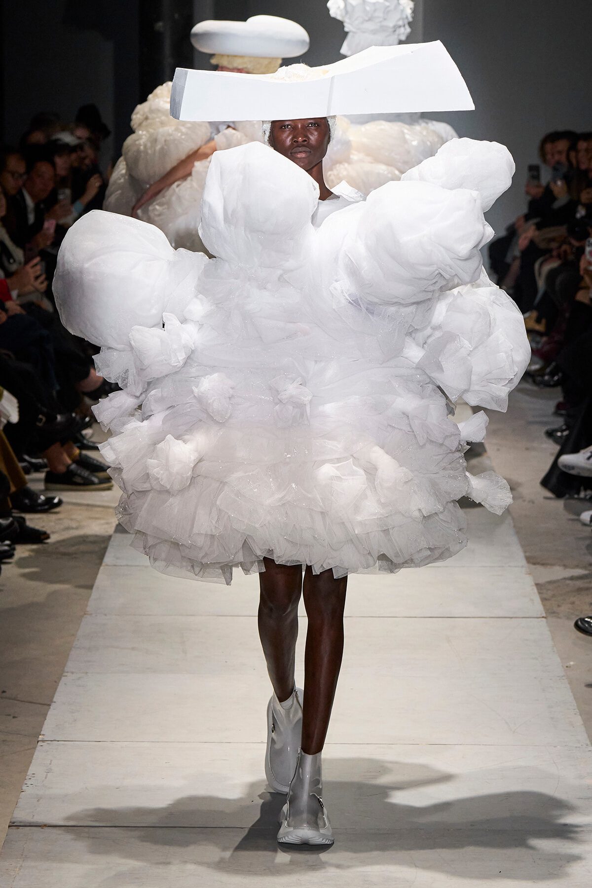 Model on a runway wearing a dramatic white sculptural dress with oversized ruffles and a wide white headpiece.