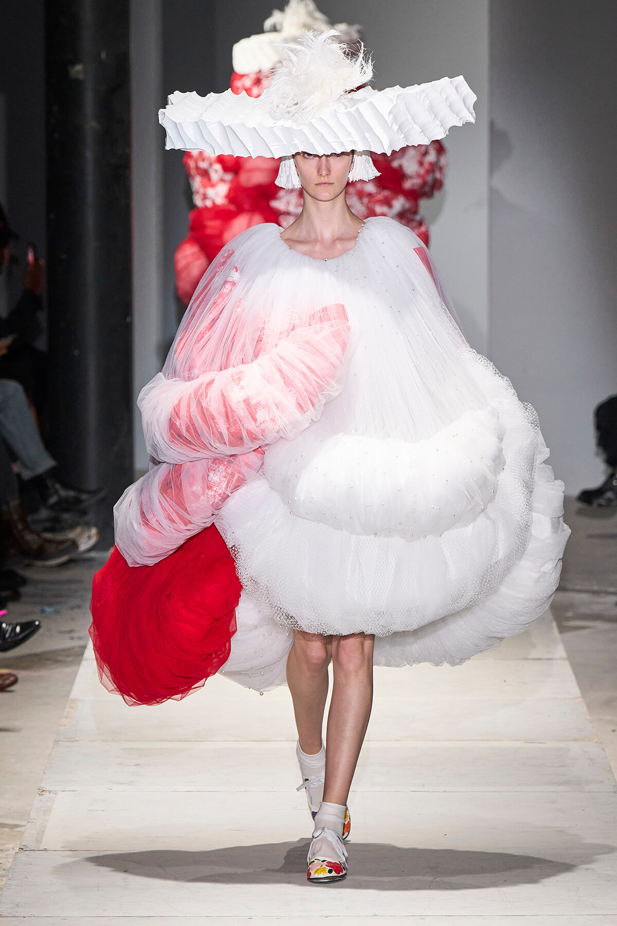 Model walks the runway in a voluminous white sculptural gown with pink underlayers and a large feathered hat.