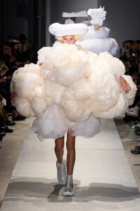 Runway model in a voluminous cream cloud-like dress with oversized puff layers, white hat, and silver boots. Audience lines the runway behind her.