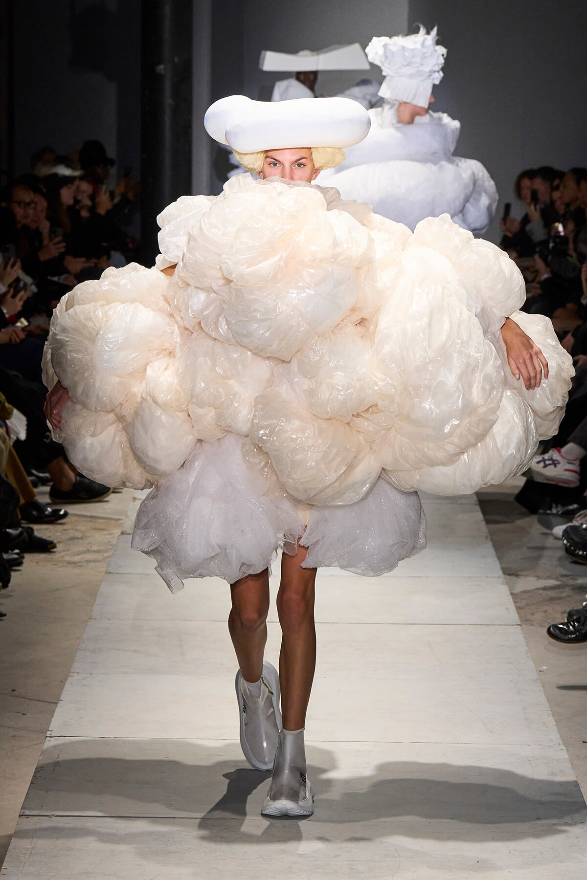 Runway model in a voluminous cream cloud-like dress with oversized puff layers, white hat, and silver boots. Audience lines the runway behind her.