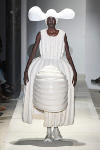 Model walks a runway in an all-white sculptural gown with a large cloud-like headpiece and rounded layered bodice and skirt.