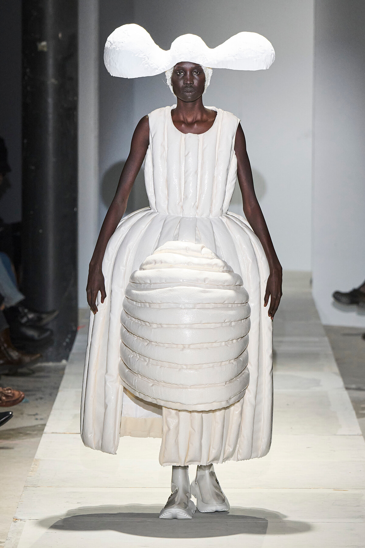 Model walks a runway in an all-white sculptural gown with a large cloud-like headpiece and rounded layered bodice and skirt.