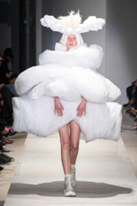 Model on a runway wearing an avant-garde white dress with cloud-like, voluminous layers and a tall fluffy headpiece.
