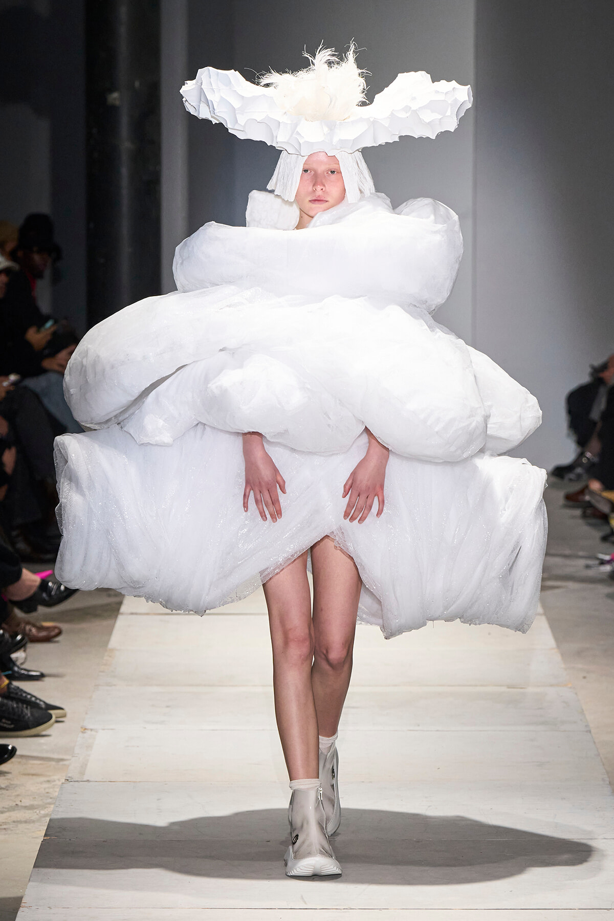Model on a runway wearing an avant-garde white dress with cloud-like, voluminous layers and a tall fluffy headpiece.