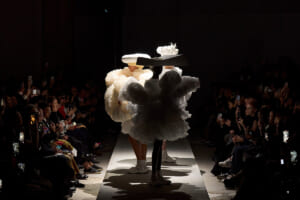 Models wear oversized puffball gowns walking a white runway while audiences on both sides film and photograph the show.