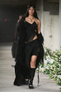 Model walks a runway in a black, asymmetrical dress with lace panels and a long feathered coat, paired with knee-high boots.